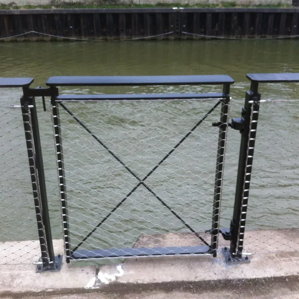 A metal gate with a crisscross wire design stands closed at the edge of a waterway, with murky green water visible beyond. The gate is part of a railing system and has a latch on the right side.