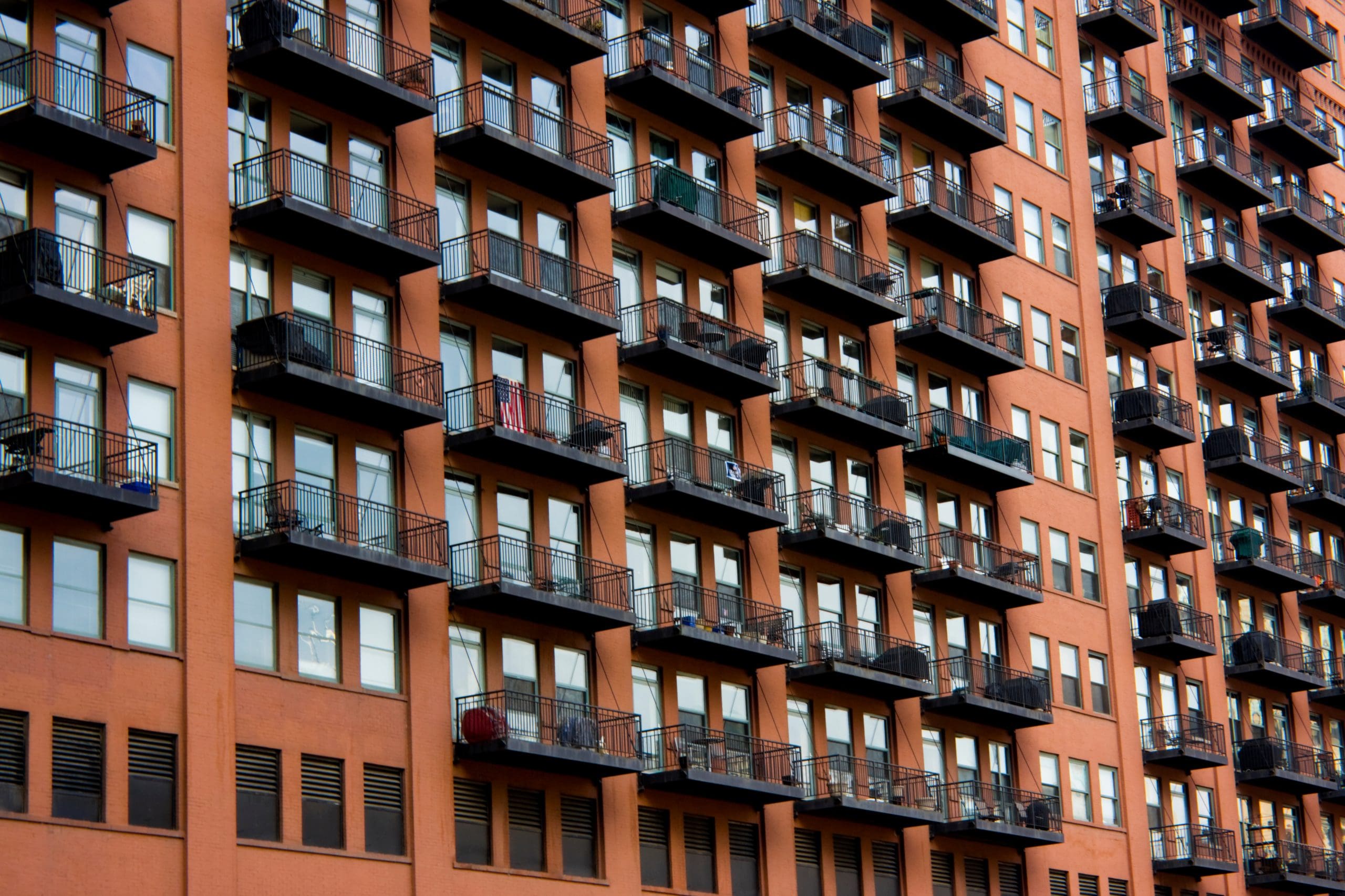 A large brick apartment building with multiple floors featuring uniform balconies and windows, creating a repeating, geometric pattern. Some balconies have plants and furniture, adding a touch of personal detail to the otherwise structured facade.