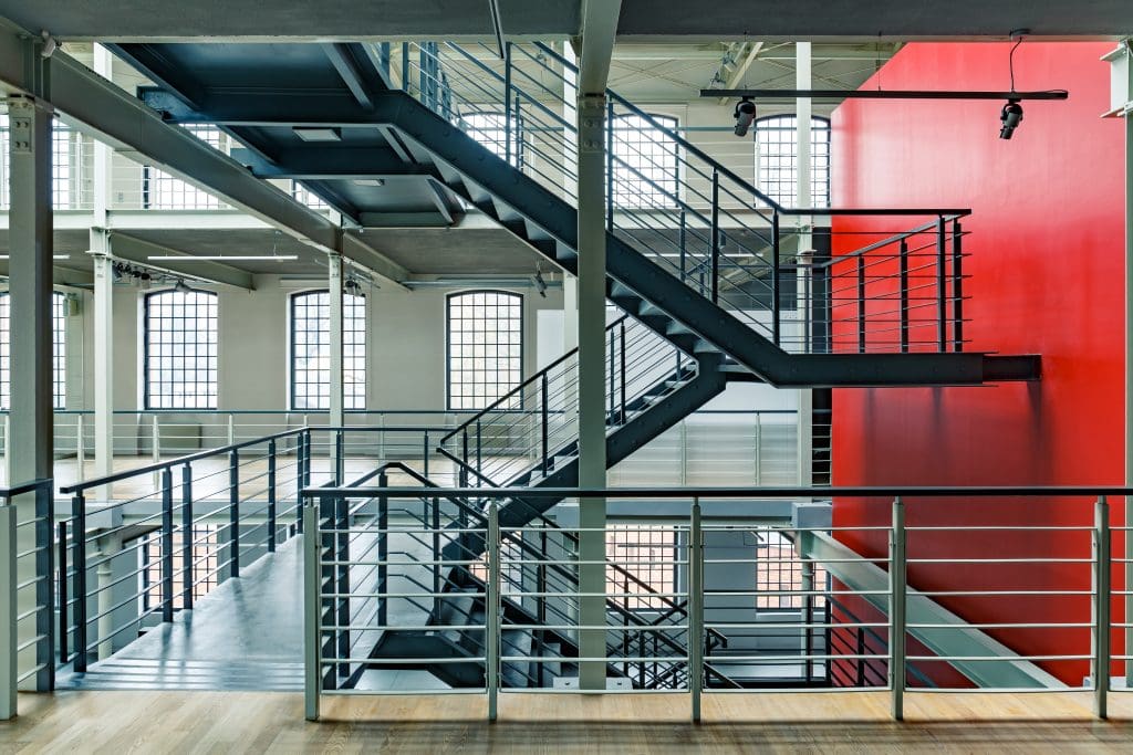 A modern industrial interior with metal staircases and railings. The space features large grid-pattern windows and a prominent red accent wall, providing contrast to the gray and metal tones of the structure.