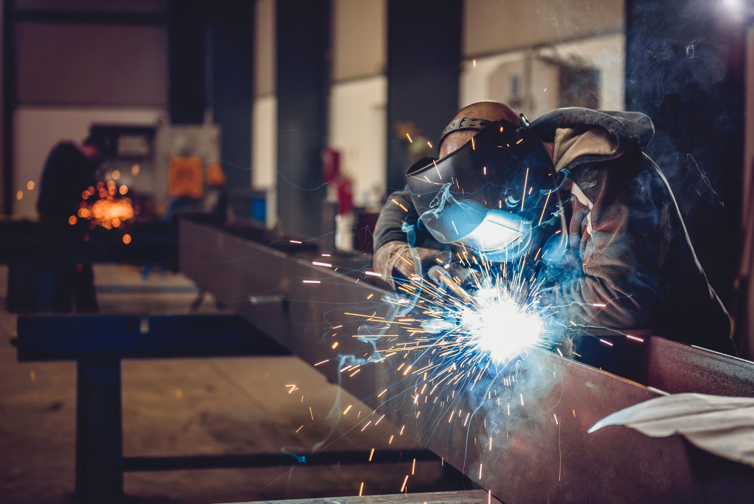 A welder wearing protective gear is welding a large metal beam in an industrial workshop. Bright sparks fly around as the welder works, illuminating the scene. The workshop appears spacious with other metal structures in the background.