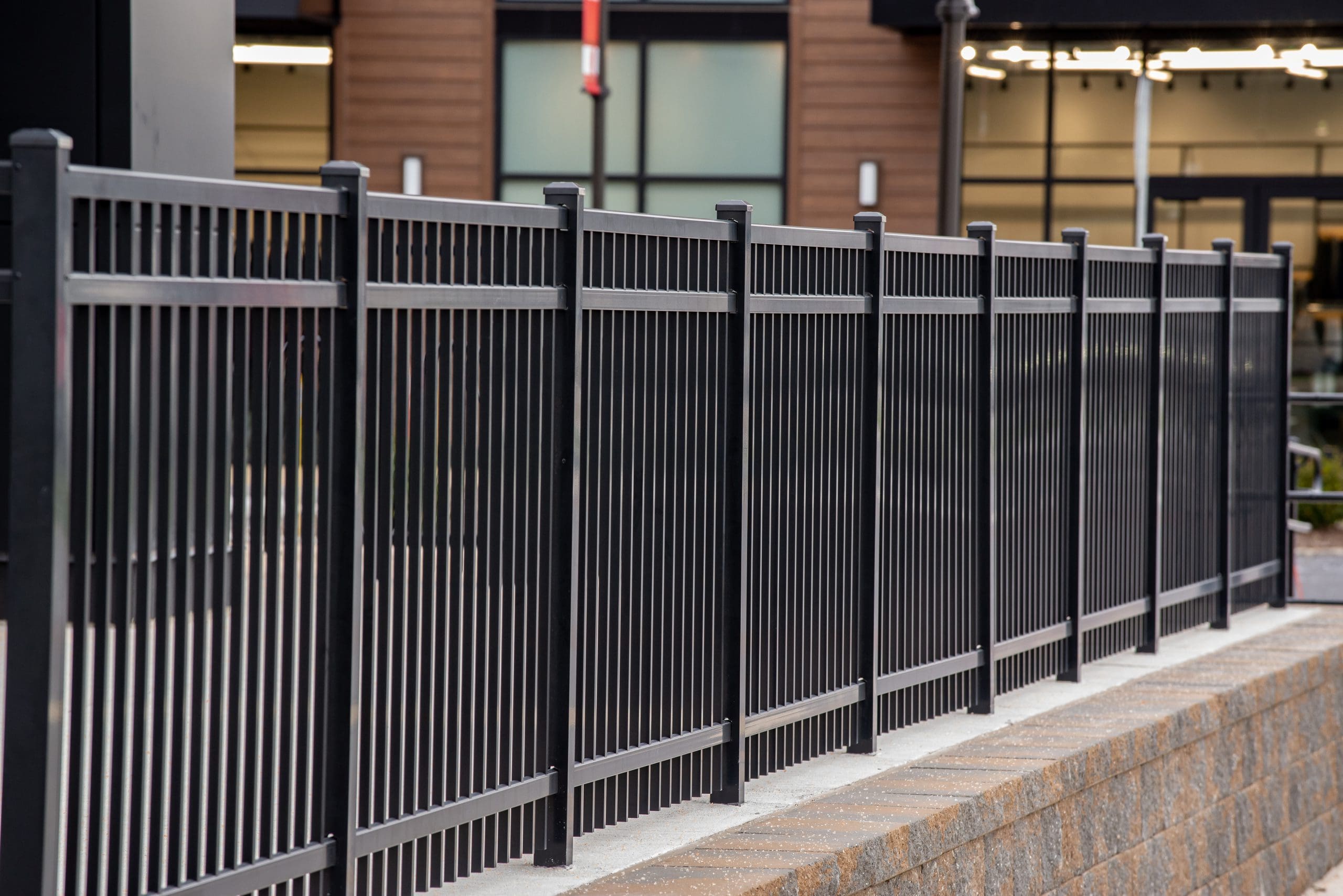 A long metal fence with vertical bars stands on a brick base in front of modern building windows. The fence stretches into the distance, creating a linear perspective.