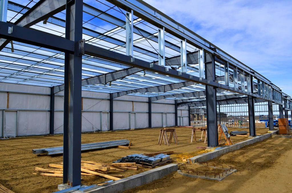 A large metal-framed structure under construction with a sandy ground. Steel beams and scaffolding are visible under a partially completed roof. The background shows a concrete wall and construction materials scattered around. Sky is partly cloudy.