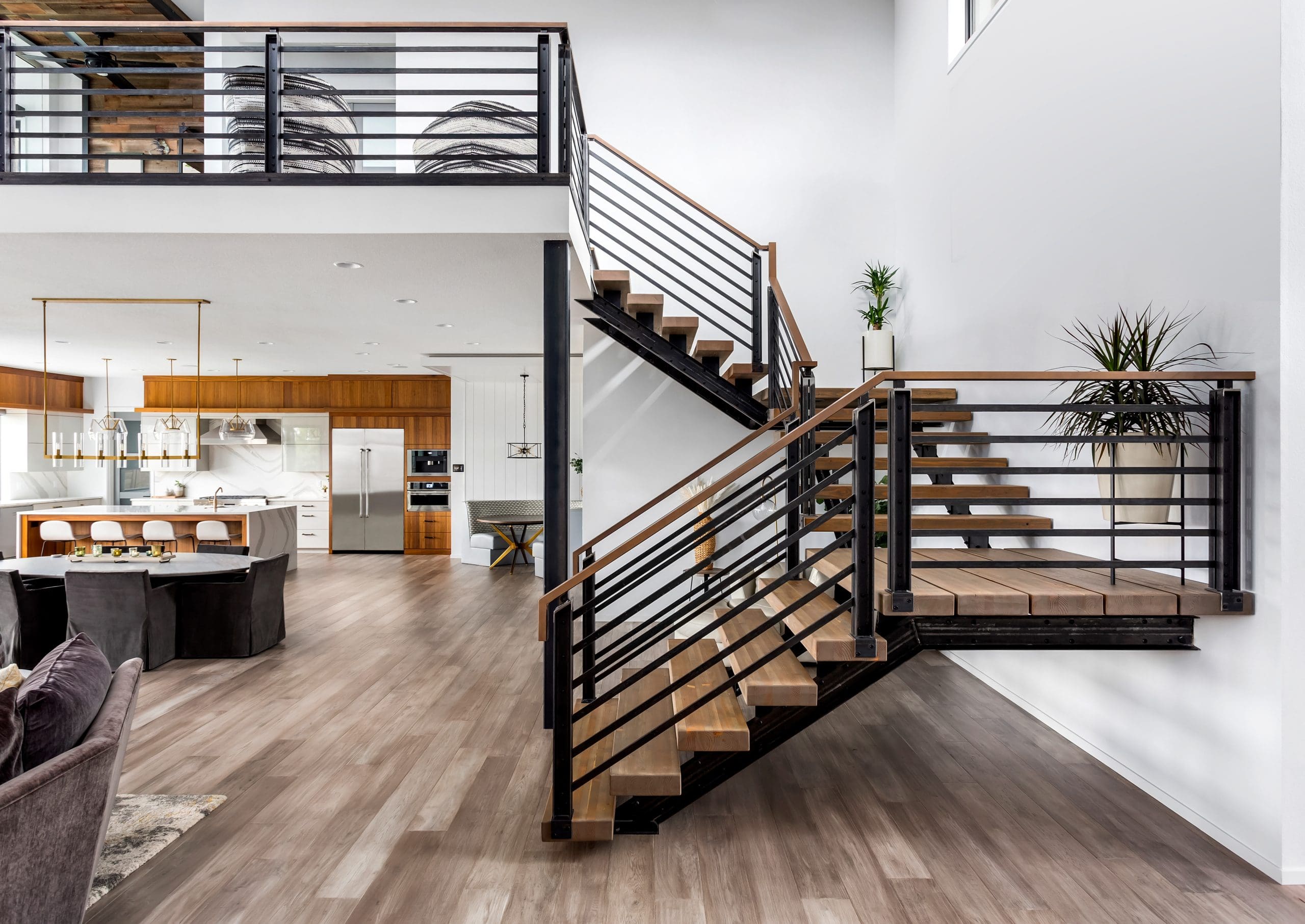 Modern interior featuring a spacious open-concept kitchen and dining area with sleek white cabinets and wooden accents. A stylish staircase with metal railings leads to the upper floor. Large windows allow natural light to enhance the space.