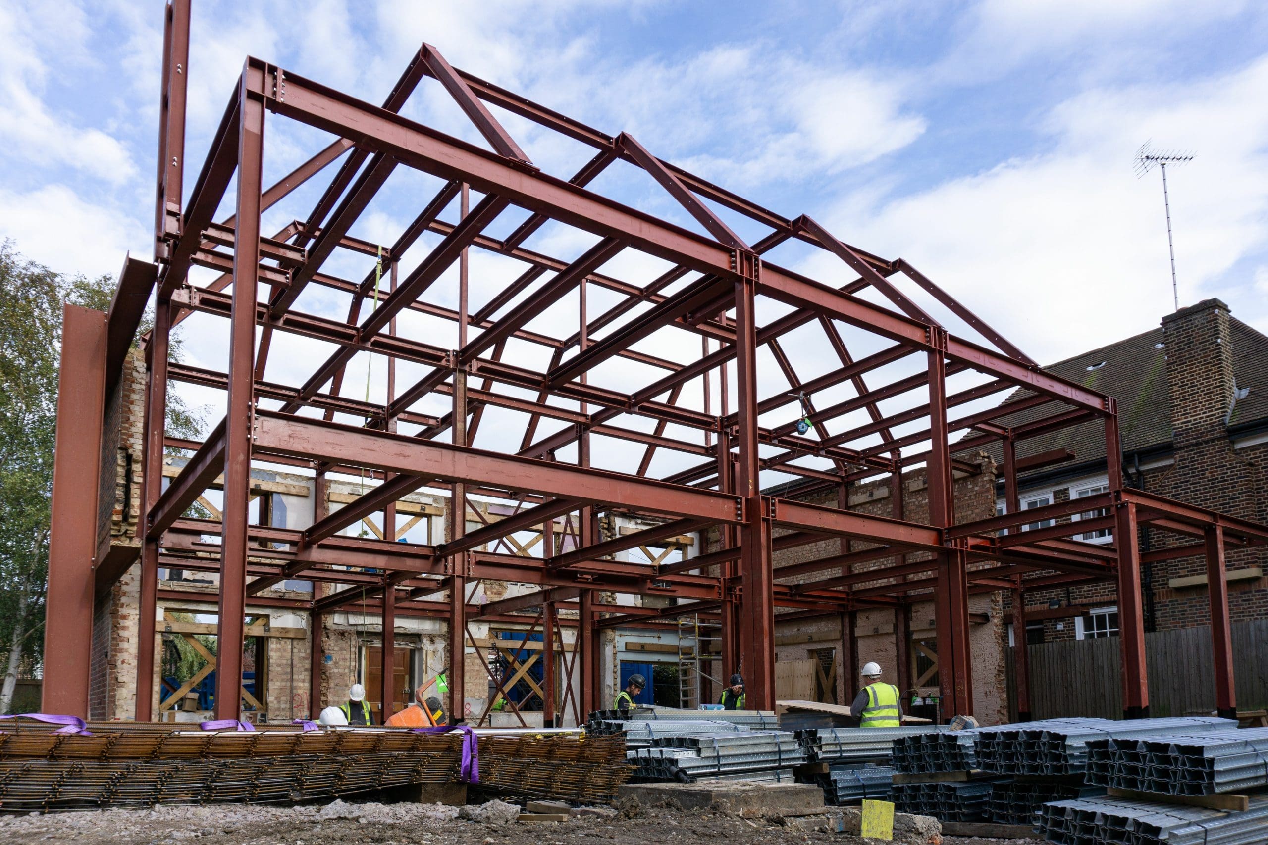 Construction site with a steel framework of a building under development. Workers wearing helmets are visible near the structure. The sky is partly cloudy, and piles of materials are scattered around the site.