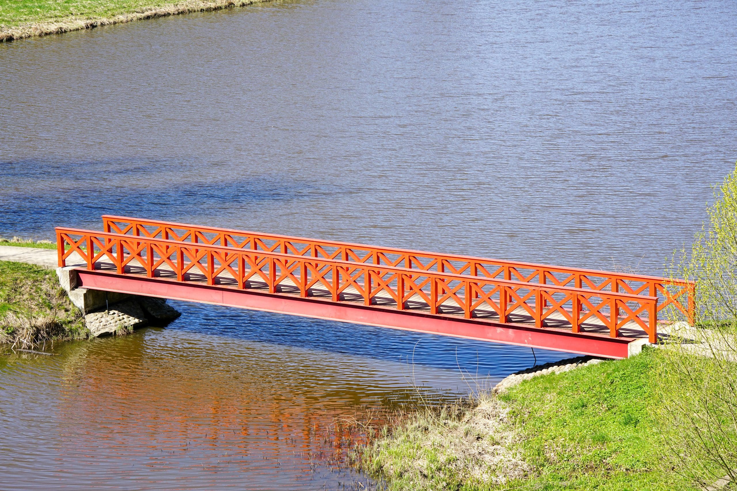 A vibrant orange pedestrian bridge spans a calm river, surrounded by grassy banks. The bright color of the bridge contrasts with the blue water, creating a picturesque landscape on a clear day.