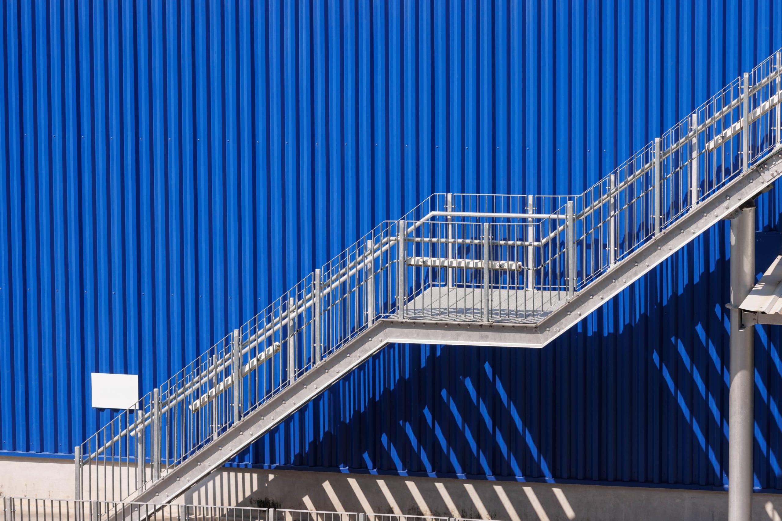 A silver metallic staircase ascends against a vibrant blue corrugated metal wall. The sun casts distinct shadow patterns on the wall, highlighting the staircase's geometric structure.