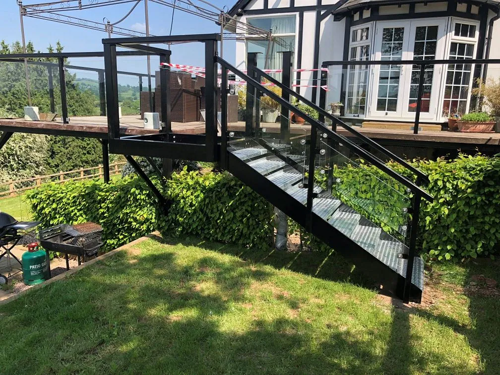 A glass and metal staircase leads from a wooden deck down to a grassy yard. The deck has a black railing and there are red and white no-entry tape sections. A house with large windows is visible in the background, surrounded by trees and shrubs.