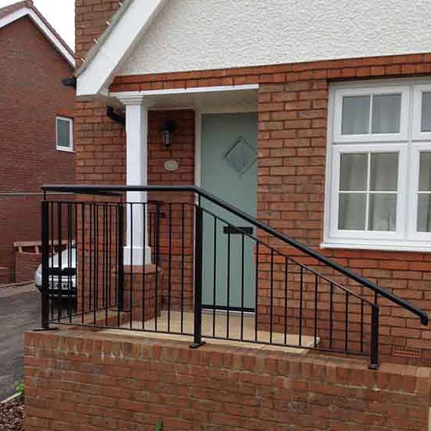 The image shows the entrance of a brick house with a light green door, a small porch, and a black metal railing along the steps. There's a window with white frames on the right and a sloped roof above. A car is partially visible in the driveway.