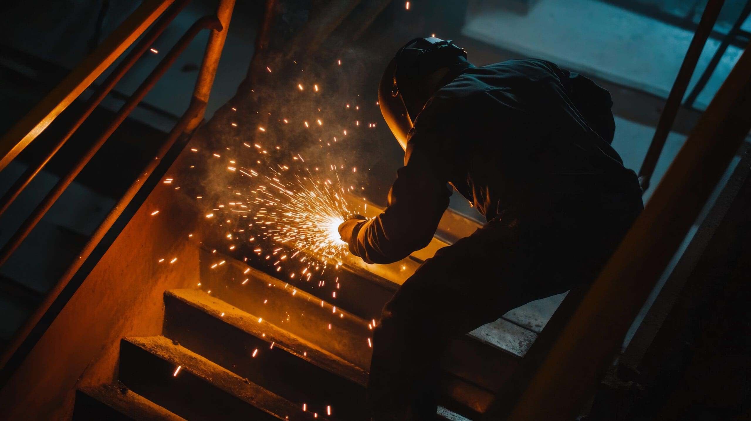 A person wearing protective gear uses a tool to create sparks on a staircase in a dimly lit industrial setting.