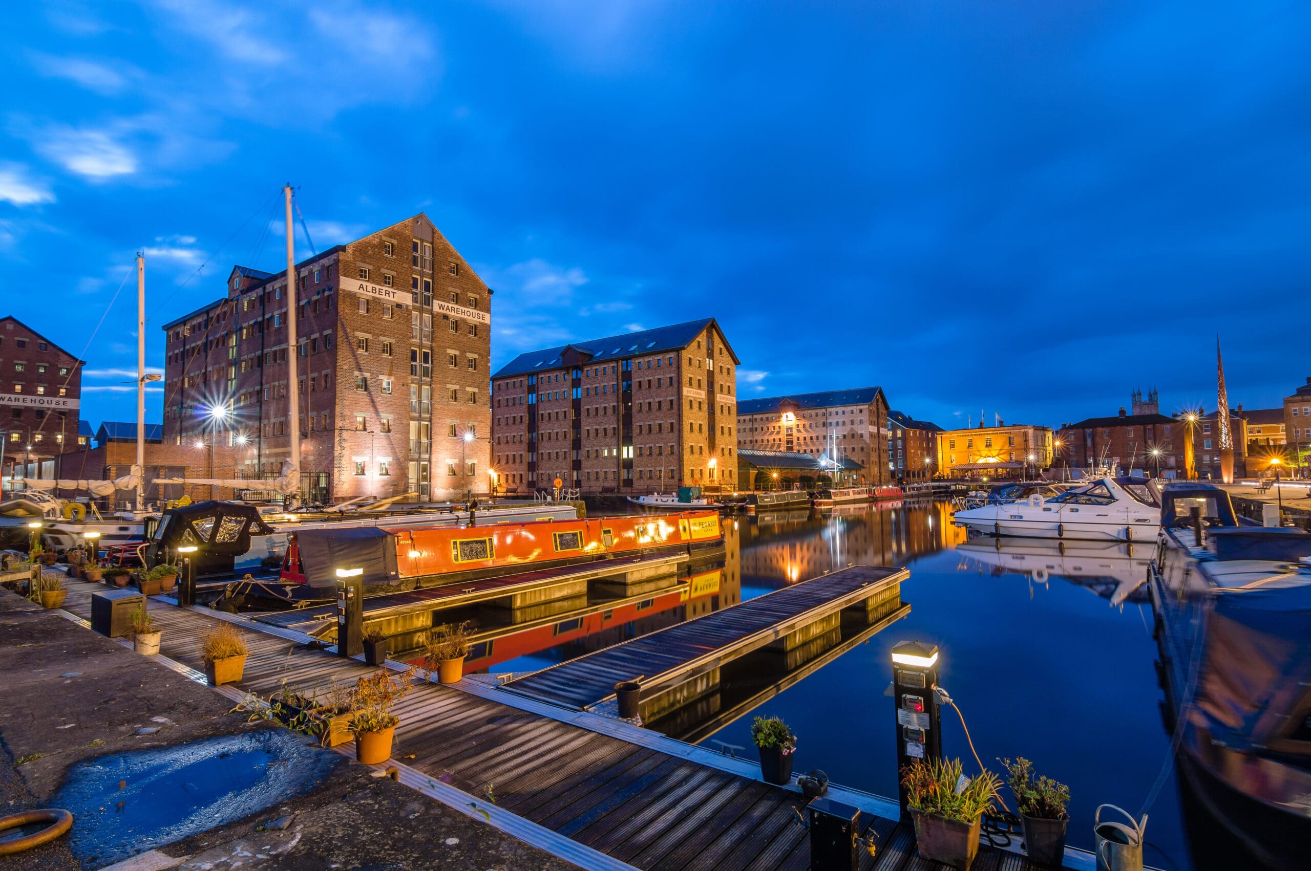 A tranquil waterfront scene at dusk, featuring brightly lit brick buildings and moored boats with their reflections visible in the calm water. The sky is a deep blue with scattered clouds.