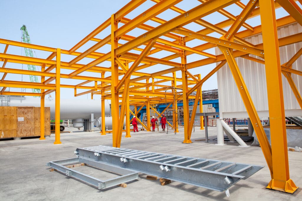 Construction site featuring a yellow steel framework structure. A metal ladder lies on the ground, and several workers are visible in the background. A crane and machinery are present, with a clear sky above.