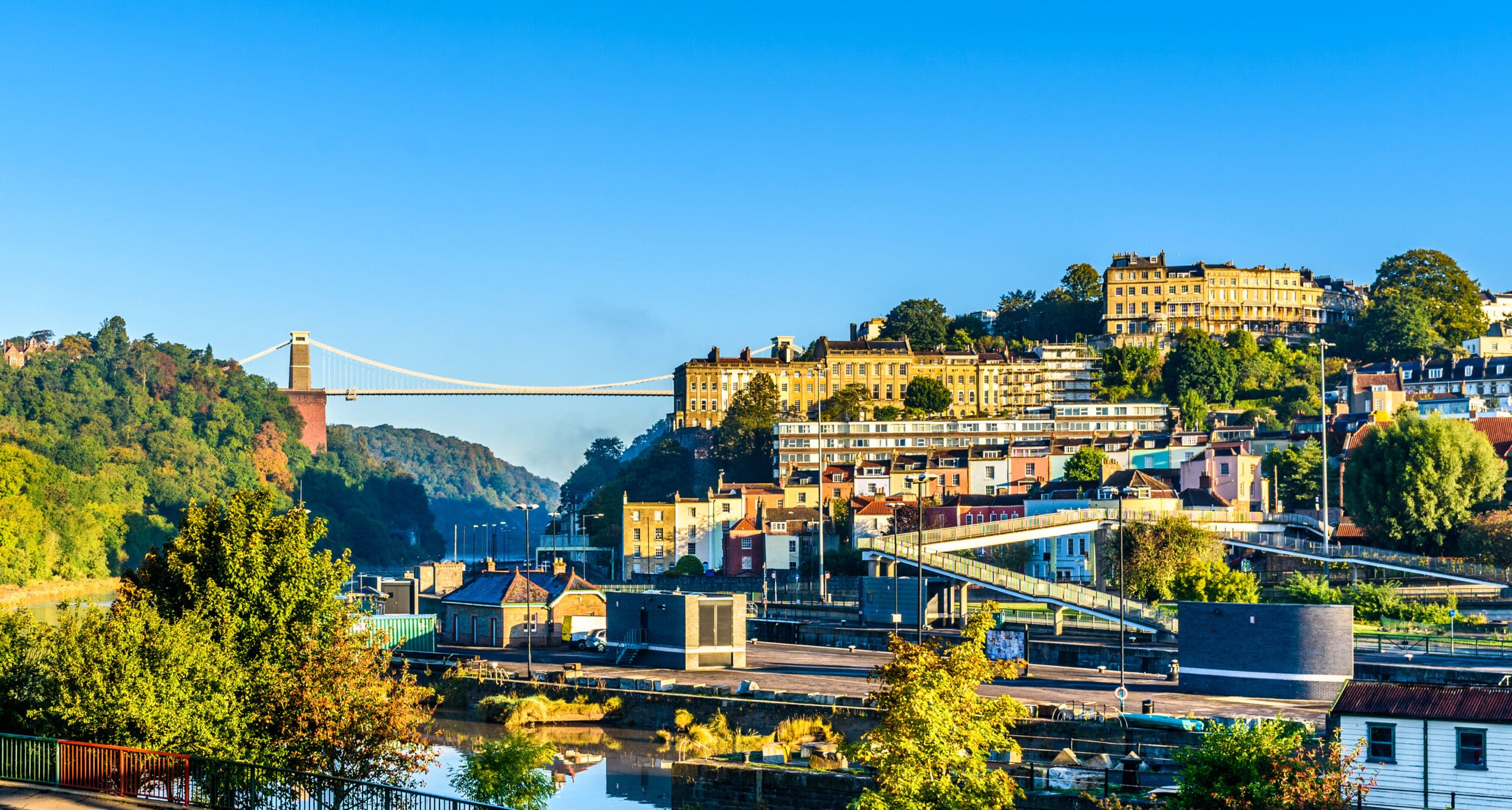 View of a sunny hillside cityscape featuring colorful houses and the Clifton Suspension Bridge spanning a lush green gorge. A calm river in the foreground reflects the vibrant scenery under a clear blue sky.