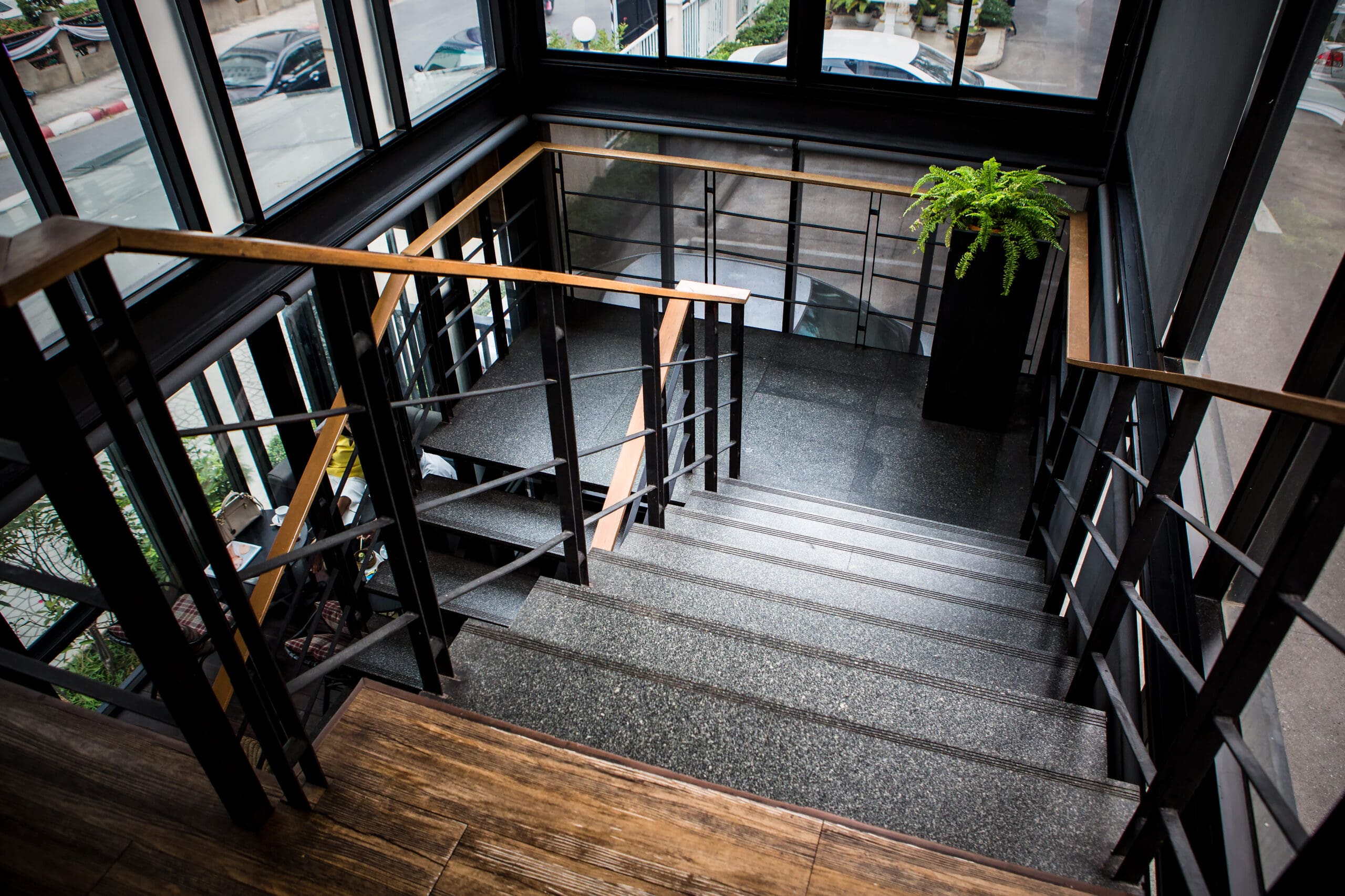 View of a modern indoor staircase with black metal railings and stone steps. A wooden handrail runs alongside. A potted fern sits at the top corner. Large windows provide natural light and a view outside.