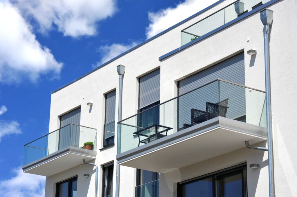 Modern apartment building with white facade and glass balconies against a blue sky with clouds. The balconies have metal railings and some outdoor furniture, including a table and chairs. A small potted plant is visible.