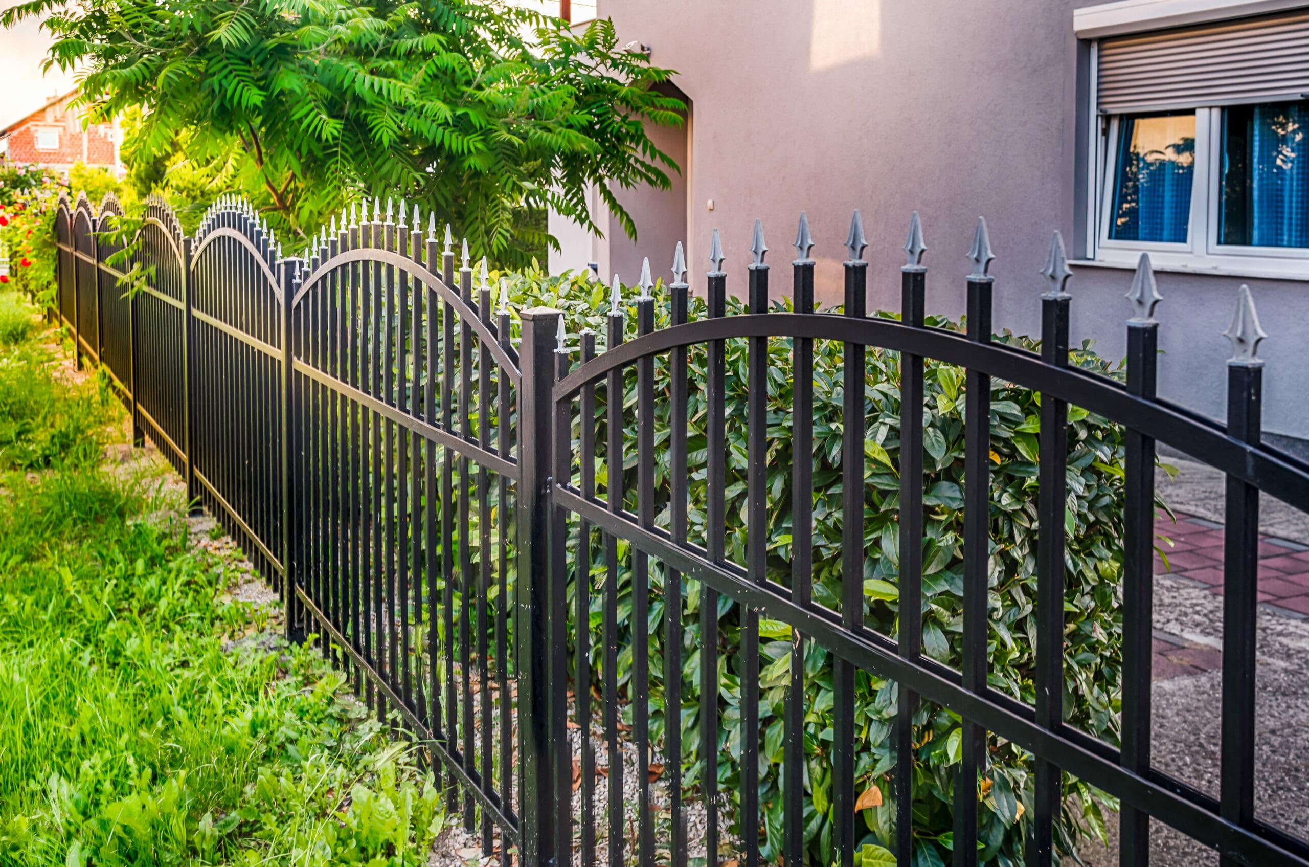 A black metal fence with pointed tops runs alongside a sidewalk next to a residential building. Green shrubs and a small tree line the fence, with sunlight creating shadows on the ground.