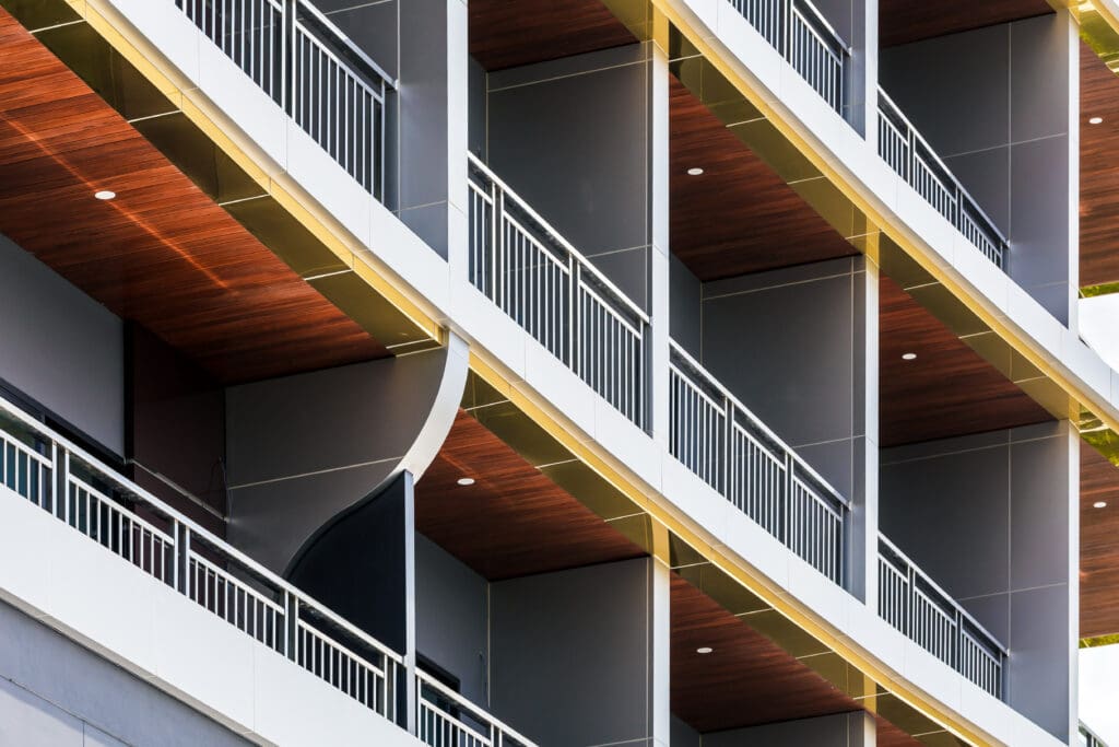 Close-up of a modern building with multiple floors featuring curved balconies and sleek metal railings. The ceilings of the balconies are lined with wood, creating a contrasting design with the building's gray and white facade.