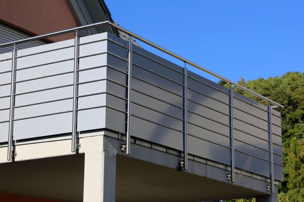 A modern balcony with sleek horizontal metal panels and a simple railing is shown against a clear blue sky. The structure is supported by sturdy pillars, with trees visible in the background.