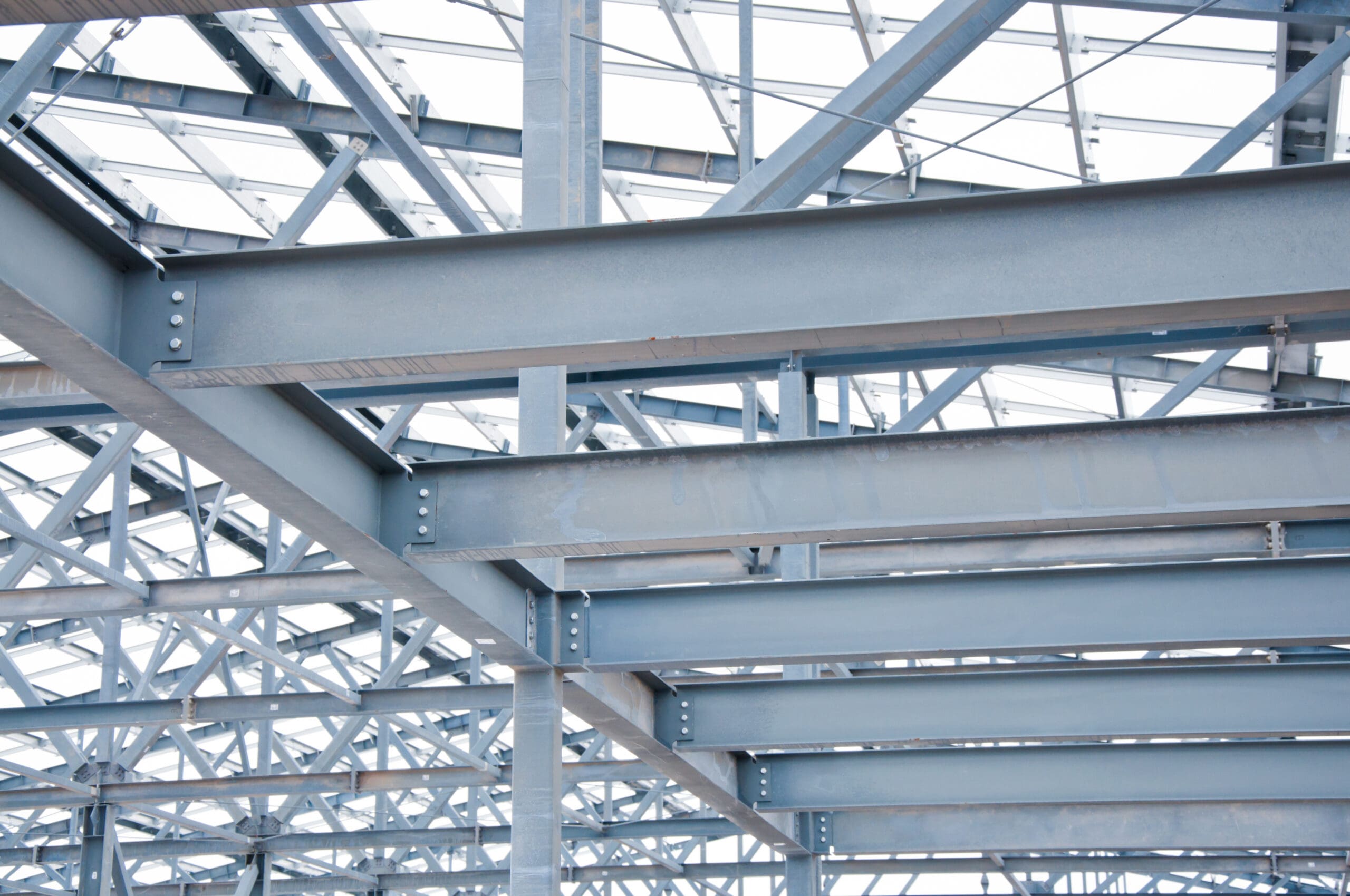 The image shows a close-up view of gray steel beams forming part of a building's frame. The beams intersect at various angles, creating a geometric pattern. The structure is set against a bright, cloudy sky.