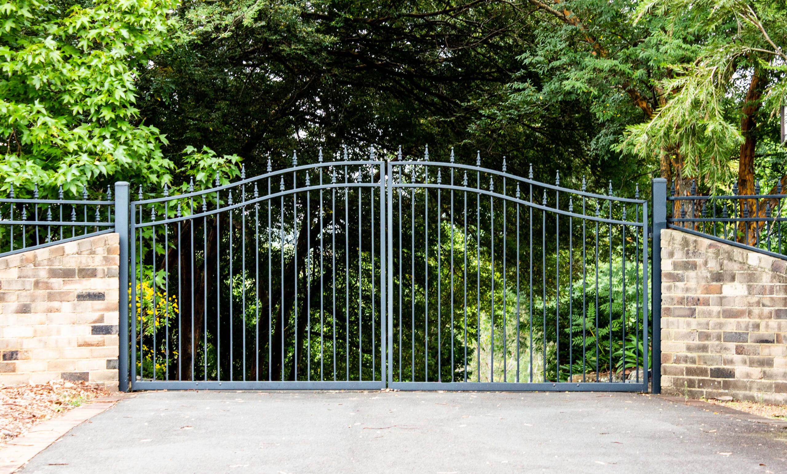 A black wrought iron gate is set between two brick pillars, leading to a driveway. The background features dense green foliage and trees, creating a secluded, woodland atmosphere.