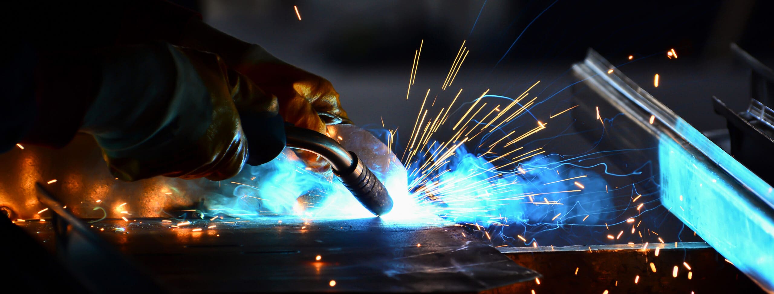 A close-up of a welder's hands in protective gloves holding a welding torch, emitting bright blue and orange sparks. The welder is working on a metal surface in a workshop, with intense light and smoke surrounding the welding area.