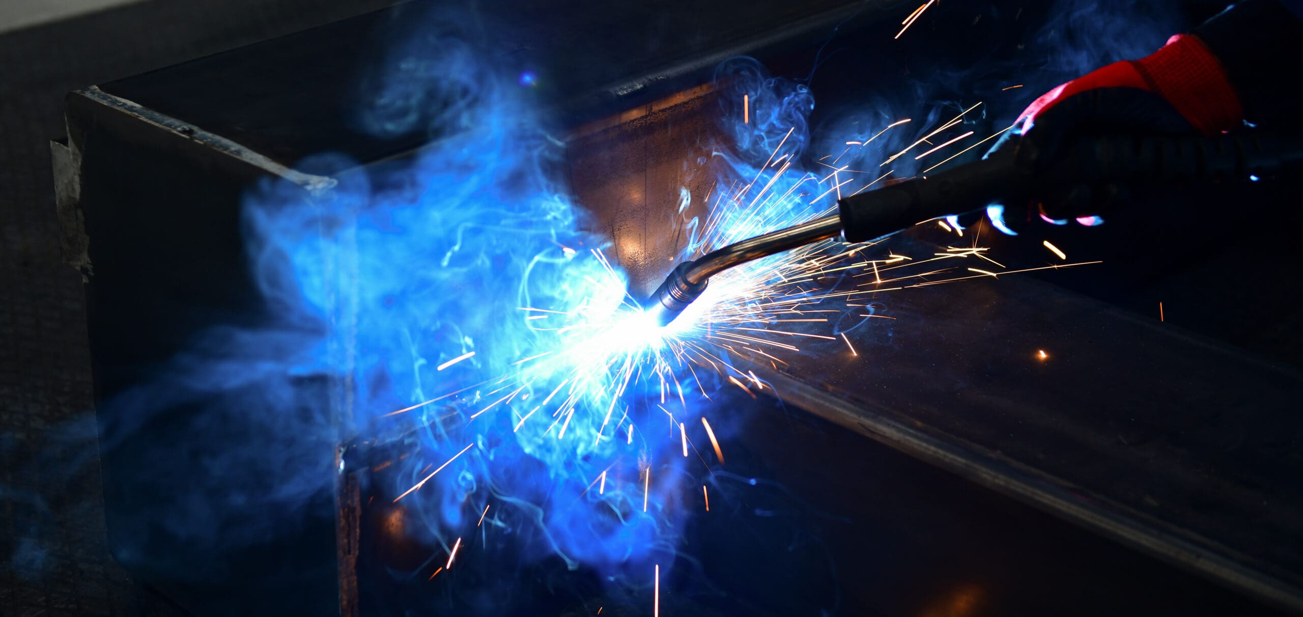 Close-up of a welding torch emitting bright sparks and blue smoke as it works on a metal surface. A gloved hand is holding the torch in a dark workshop setting.