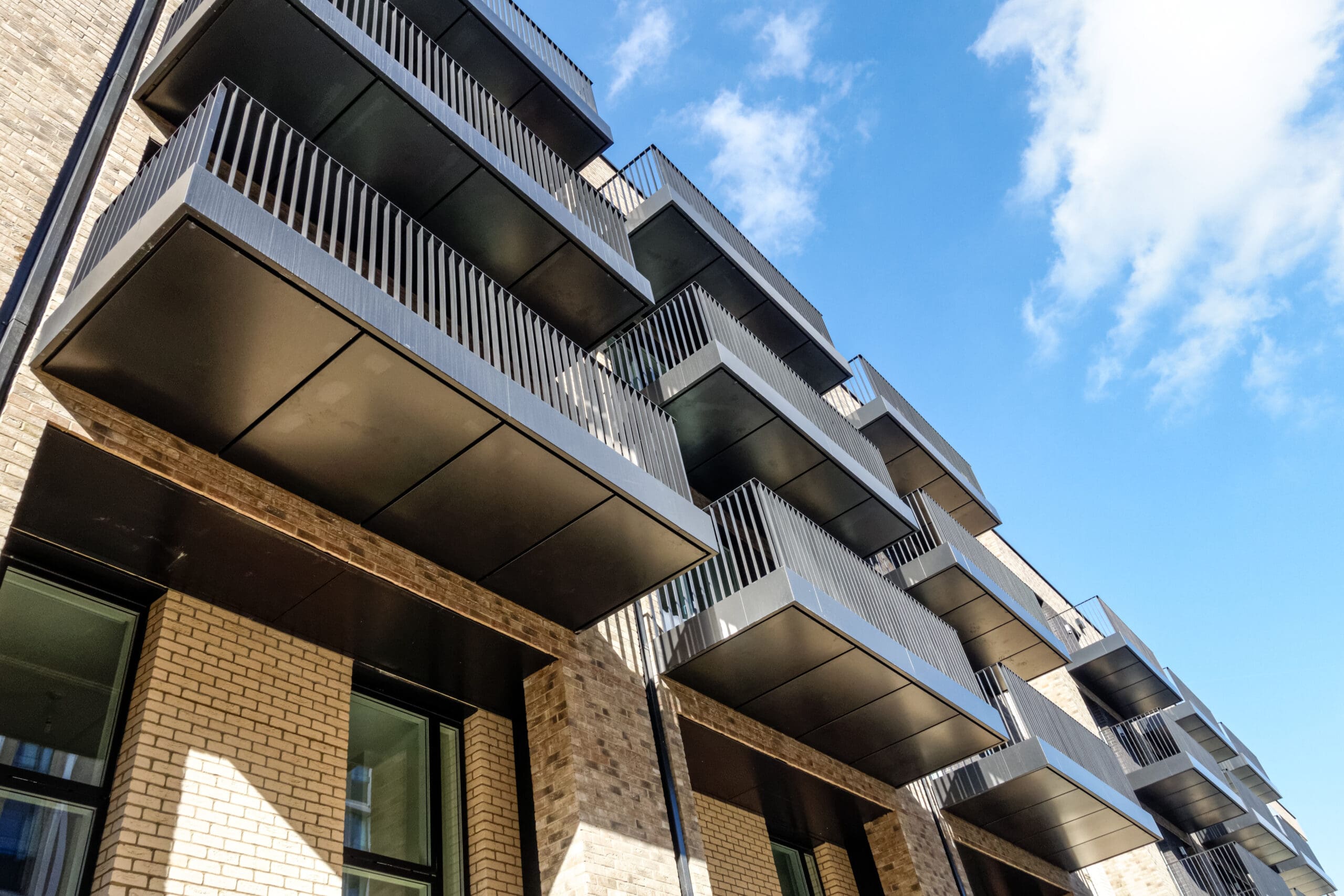 A modern apartment building with multiple balconies is shown against a blue sky. The structure features light brickwork and sleek, black metal railings.