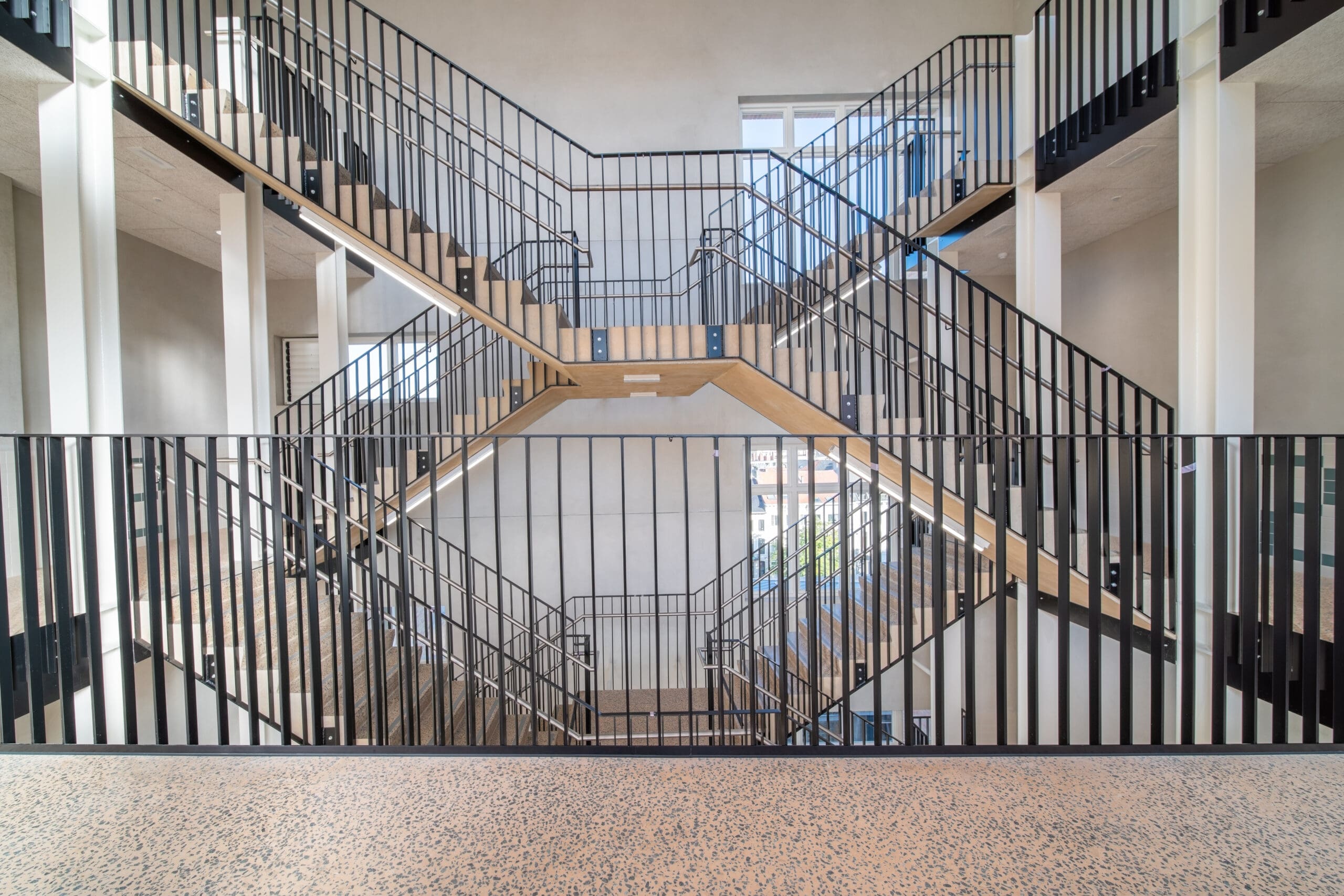 A modern, multi-level interior staircase with wooden steps and black metal railings is shown. The design features open space and natural lighting from windows, creating a bright and airy atmosphere.