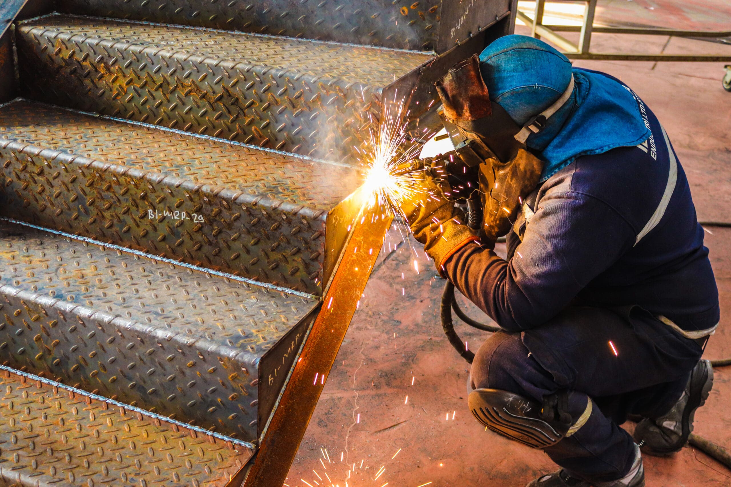 A welder in protective gear crouches while welding a metal staircase. Sparks fly as they work in an industrial setting, highlighting the intense focus and skill required for the task.