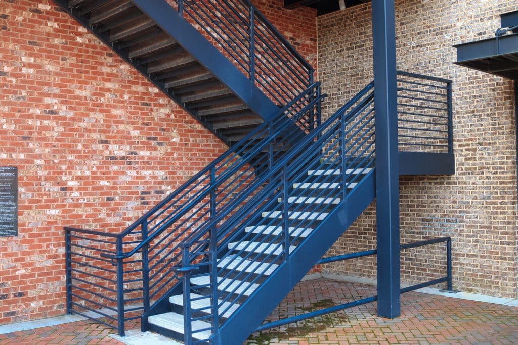 A blue metal staircase with railings is attached to a red brick wall. The stairs lead upwards in two sections with a landing in between. The ground is paved with red and tan bricks in a herringbone pattern.