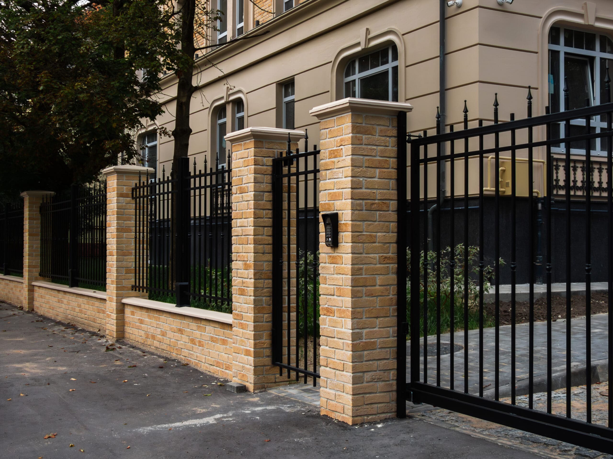 A black metal fence with brick pillars surrounds a beige building. The entrance features an automated gate with key access, adjacent to a concrete sidewalk and leaf-covered area.