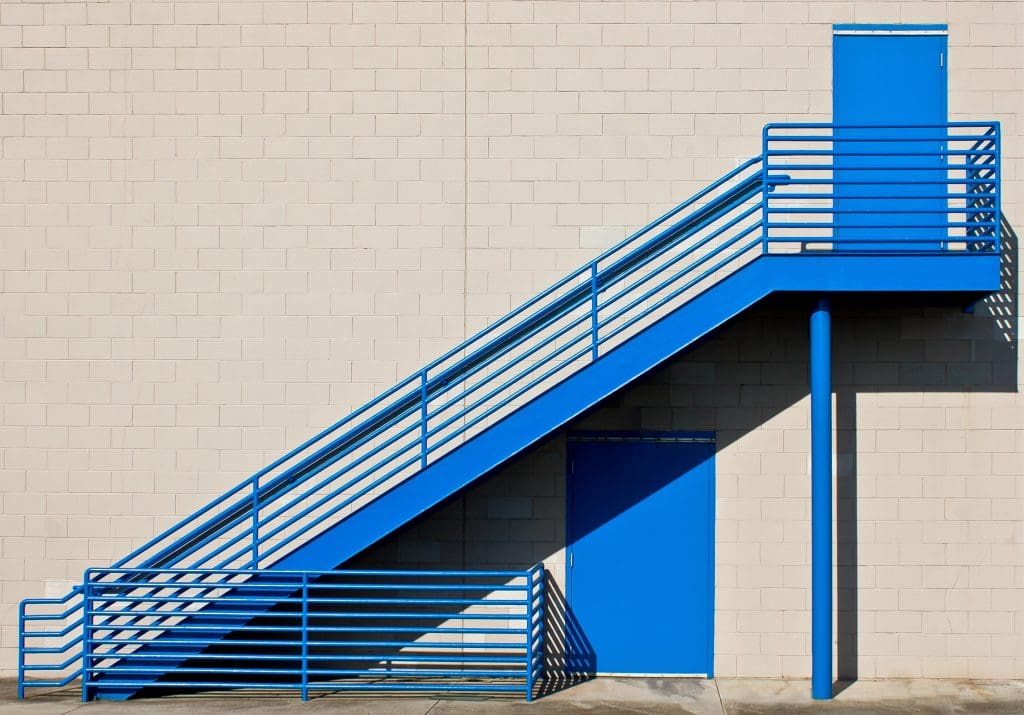 A blue metal staircase leads to a blue door on a beige brick wall. Another blue door is visible at the bottom, aligned with the staircase. The composition is simple, with strong geometric lines and contrasting colors.