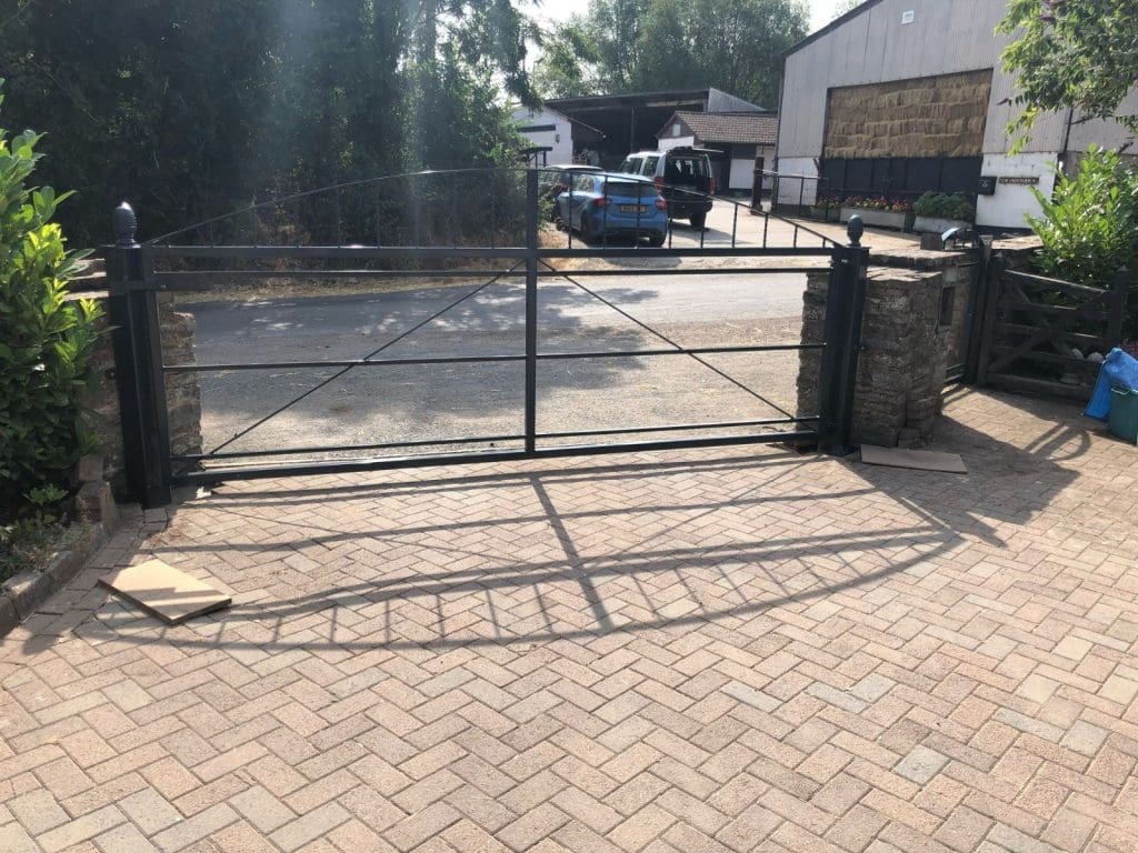 A black metal gate stands open on a paved driveway, leading to a rural property with a barn and a blue car. Trees and greenery surround the area, and sunlight casts shadows on the ground.