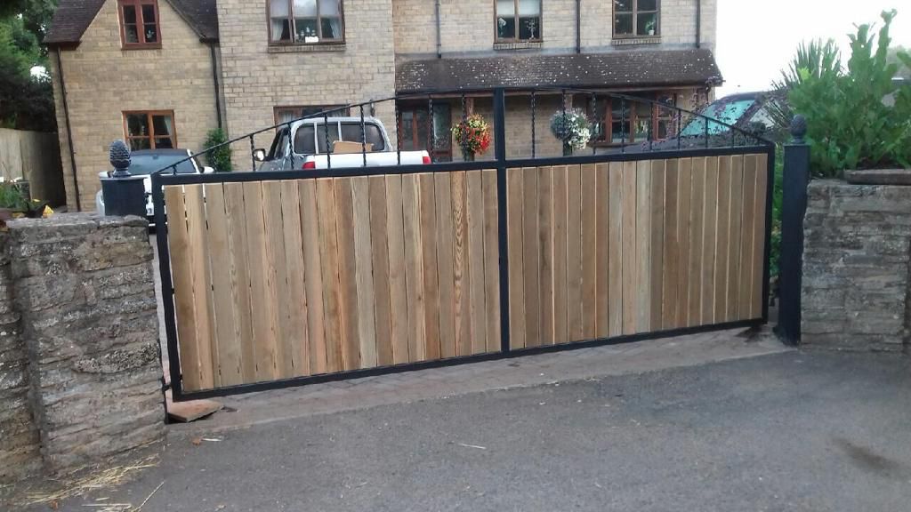 A large wooden gate with vertical planks is set between stone pillars, partially open, in front of a two-story stone house. A white truck is parked behind the gate, and potted plants are visible on the gate's edge.