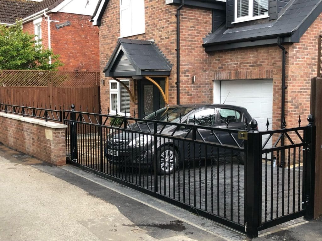 A black car is parked in the driveway of a brick house with a black-framed garage door. The driveway is enclosed by a black metal gate with pointed tops. A wooden fence runs along the side, and there is a small awning over the front door.