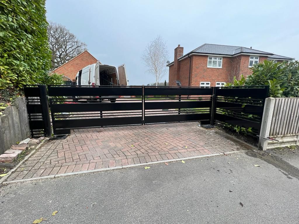 A black metal gate with horizontal slats opens onto a paved driveway leading to a modern brick house. A delivery van is parked in front of the house. Nearby, lush green bushes and trees border the property.