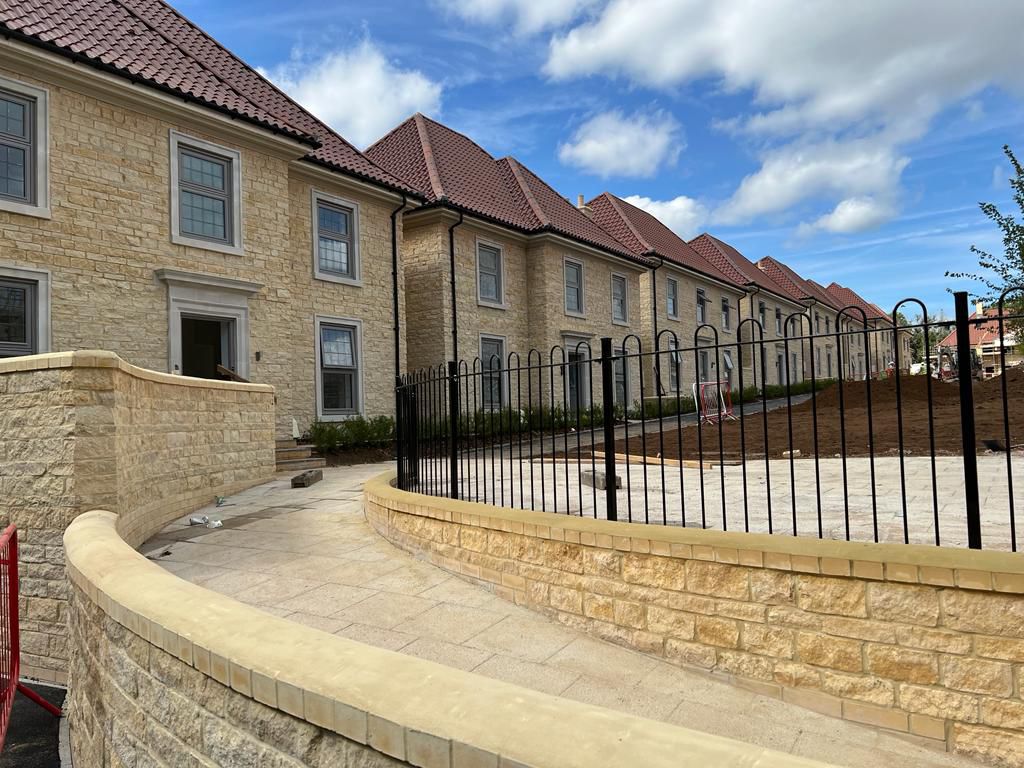 A row of newly constructed stone townhouses with red-tiled roofs. A curved pathway with a metal fence leads to the entrance. The sky is partly cloudy, and the area is landscaped with soil prepared for planting.