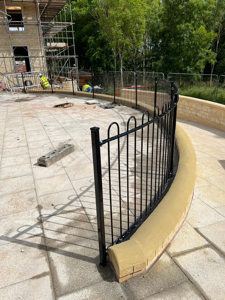 A curved metal fence lines a paved area near a building under construction. Scaffolding and construction materials are visible, along with workers and surrounding greenery.