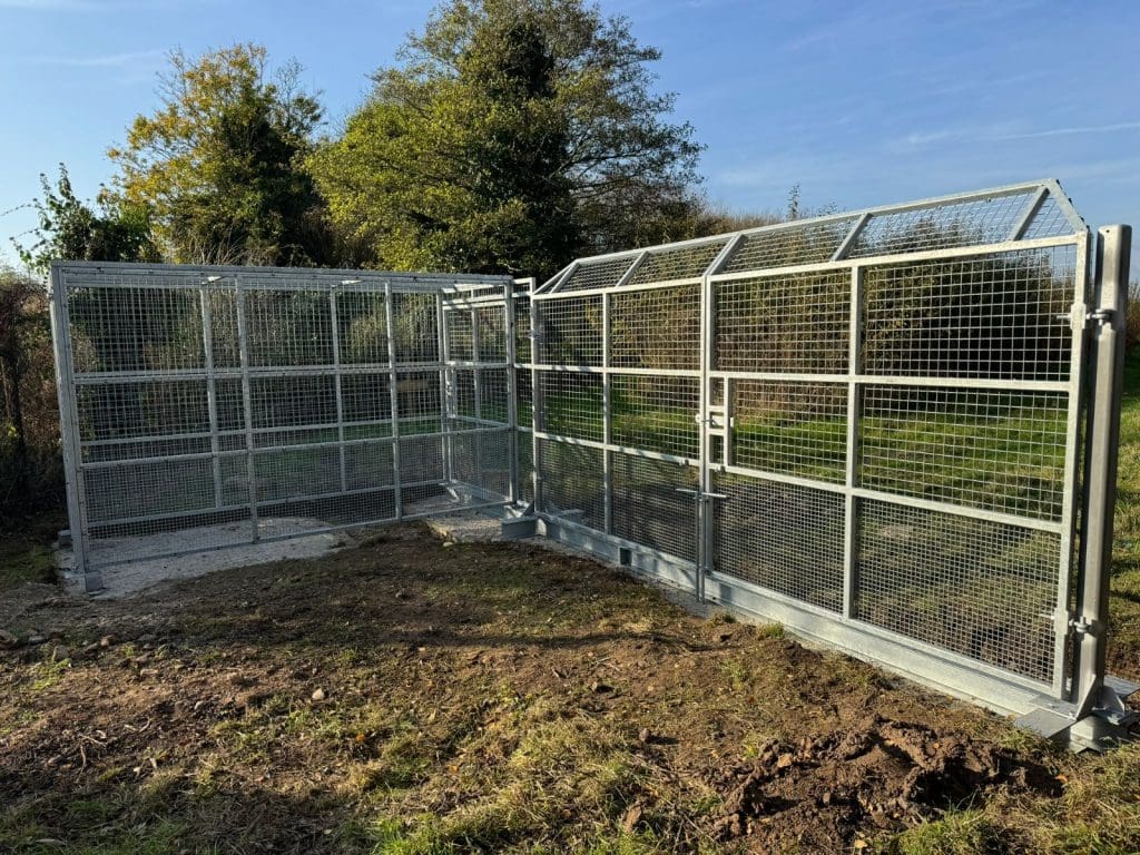 A metal enclosure with mesh fencing and a gate, situated on a grassy area with trees in the background under a clear blue sky.