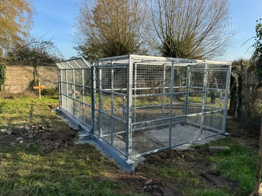 A large metal animal enclosure with a mesh roof is set on concrete in a grassy area. Trees without leaves stand behind it, and a wooden fence is partially visible in the background.