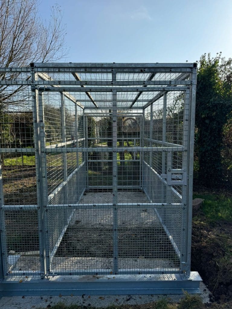 A metal cage structure stands outdoors on a concrete base, surrounded by grass and trees. The cage has wire mesh walls and a clear sky is visible above, suggesting a sunny day.