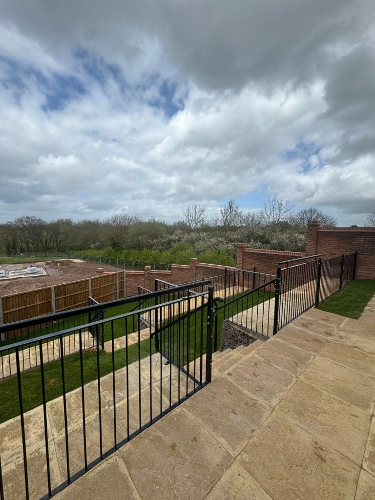 A residential backyard with a stone paved walkway and metal railing overlooking descending layers of grass and fences. The sky is cloudy with patches of blue, and trees line the horizon in the distance.