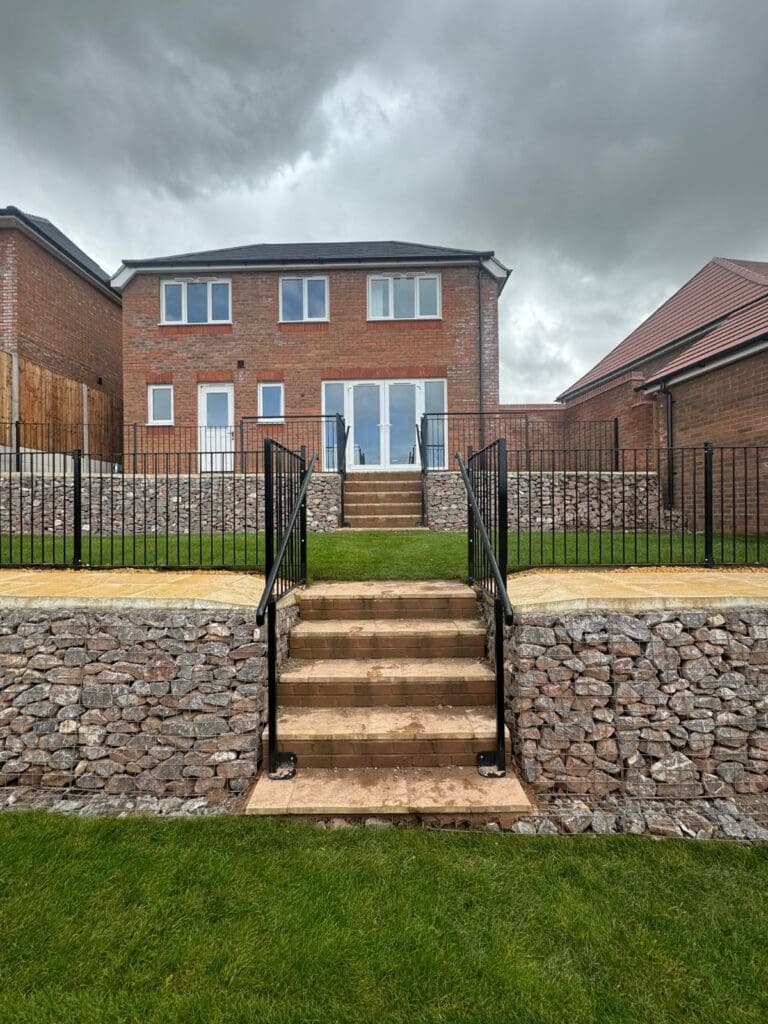 A red brick two-story house with large windows and glass doors, set on a raised stone terrace. Stone steps lead up to the house, flanked by grassy areas and metal railings. The sky is overcast.