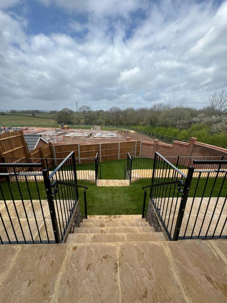 View from stone steps leading to a grassy area and fenced construction site. The scene is surrounded by trees under a cloudy sky. Black railings line the steps, and wooden fences enclose the construction area.