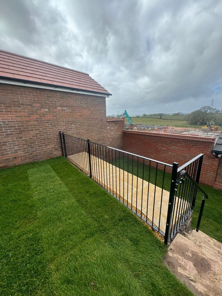 A wooden ramp with black metal railings leads from a brick house. Green grass surrounds the area, with a construction site and open field visible in the background under a cloudy sky.