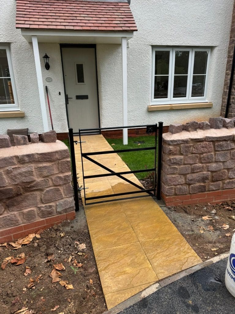 A small black metal gate opens to a path leading to a white house with a red-tiled roof and two windows. The path is lined with grass on both sides and is flanked by brick walls. Leaves are scattered on the ground.