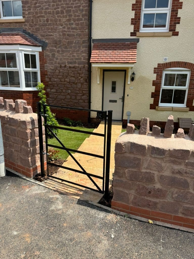 A small brick house entrance with a metal gate opens to a pathway leading to a front door. The exterior features a mix of red and cream brickwork, with small garden beds and a few plants beside the path.