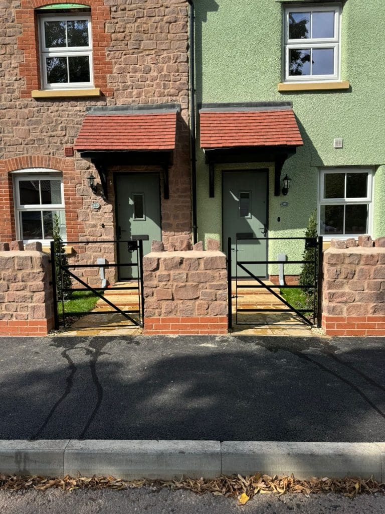 Two adjacent houses with small front gardens, each having a gate and brick pathway leading to the front doors. One house has green walls, and the other has brown brick walls. Both roofs have reddish-brown tiles.