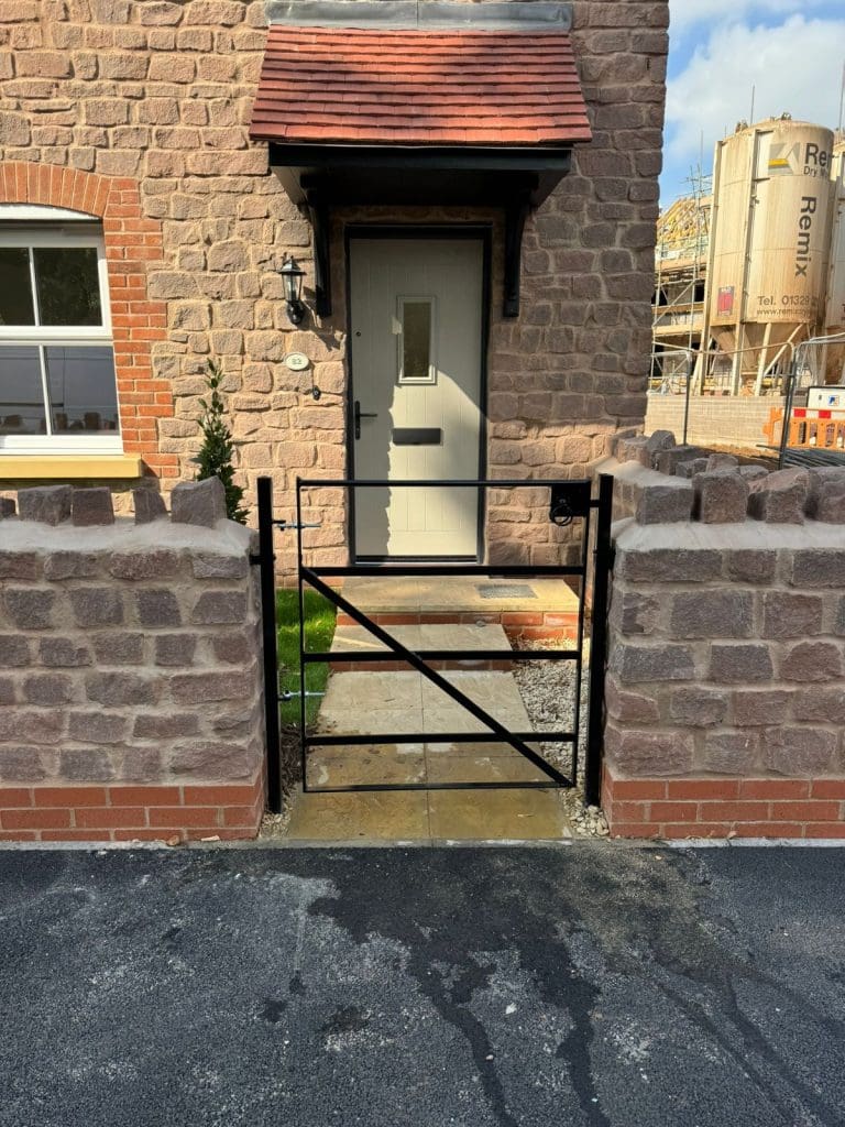 Front entrance of a house with a beige door, black metal gate, and stone walls. There's a small pathway leading to the door. A brick chimney is visible, with a construction site in the background.