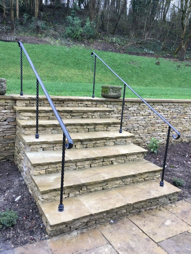 Stone steps with black metal handrails lead up to a landscaped area with green grass and trees in the background. The steps are bordered by stone walls on each side.