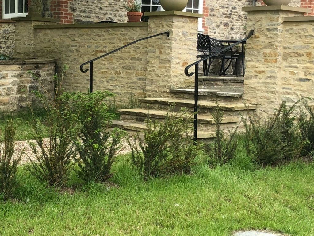 A stone staircase with black metal railings leads up to a brick and stone patio with outdoor furniture. The foreground features a grassy area with small shrubs, and a brick building with a small potted plant is in the background.